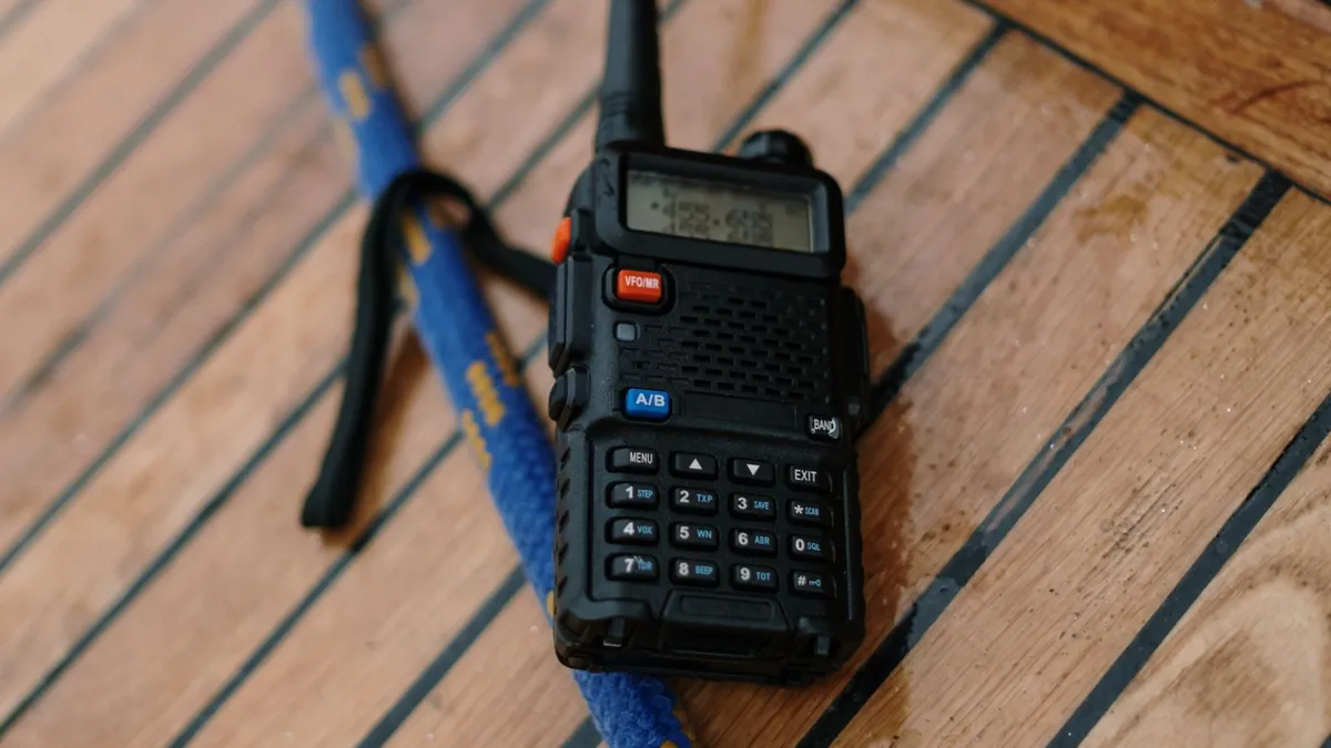 A handheld two-way radio with a long whip antenna lying on a wooden table beside a notebook