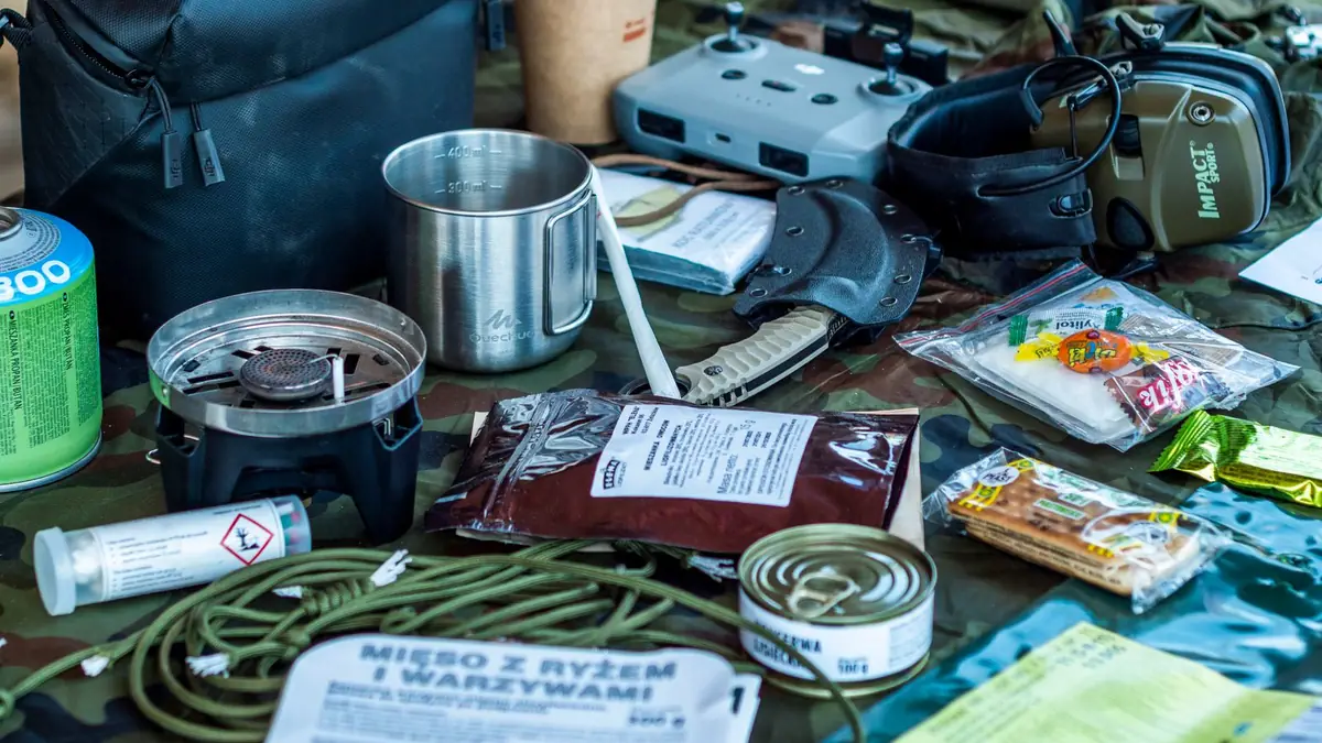 Ration pouches, a canned meal, a camp stove and a steel cup spread out on a camouflage tarp