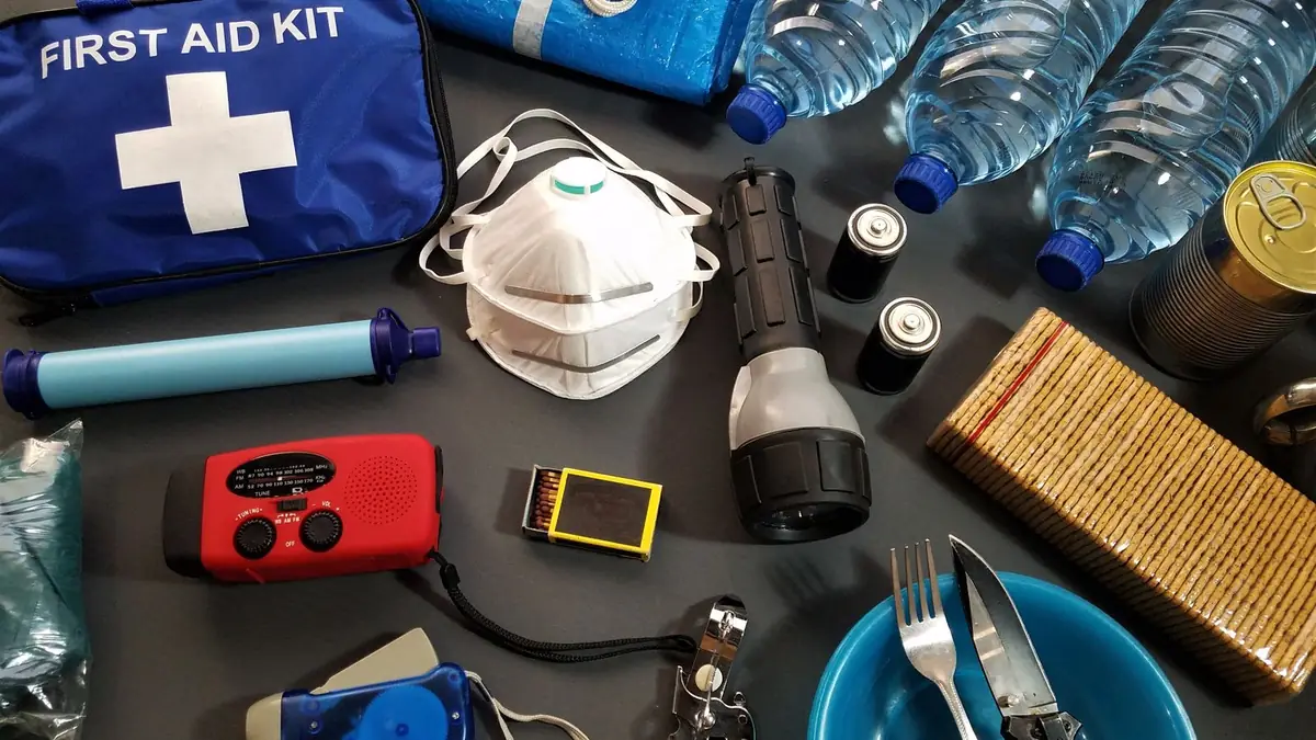 An emergency kit laid out on a dark table: first aid bag, crank radio, flashlight, batteries, water bottles and a dust mask
